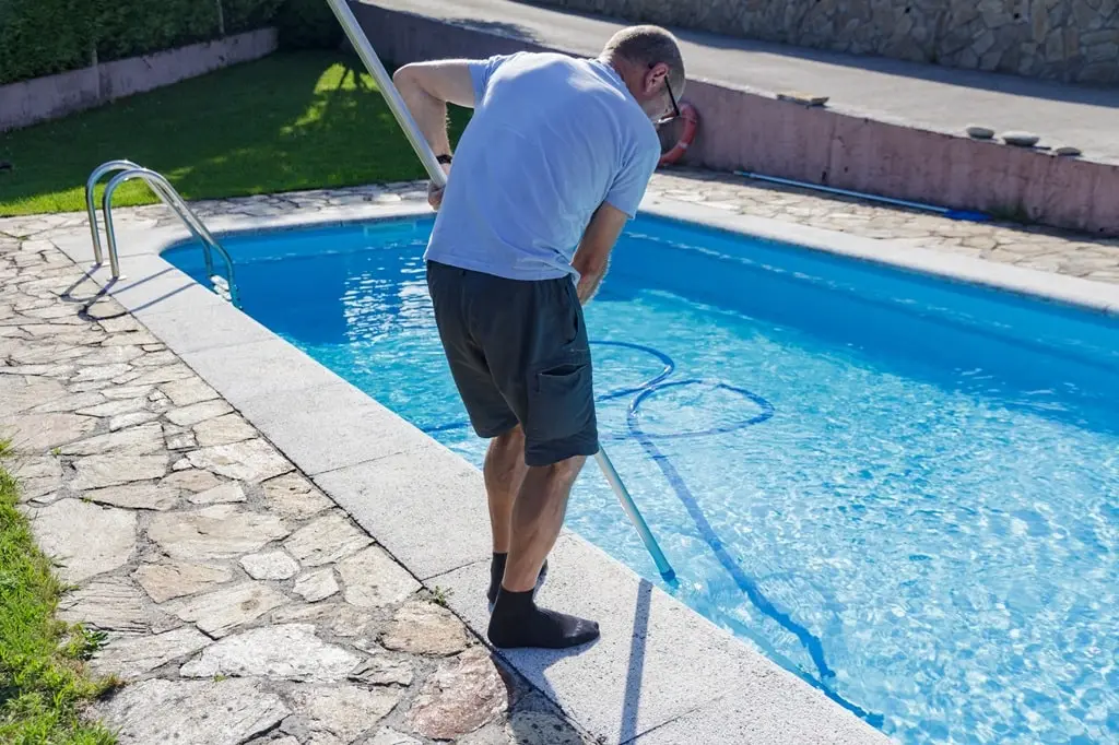 A man stands by the poolside, cleaning it with a net with a long pole.